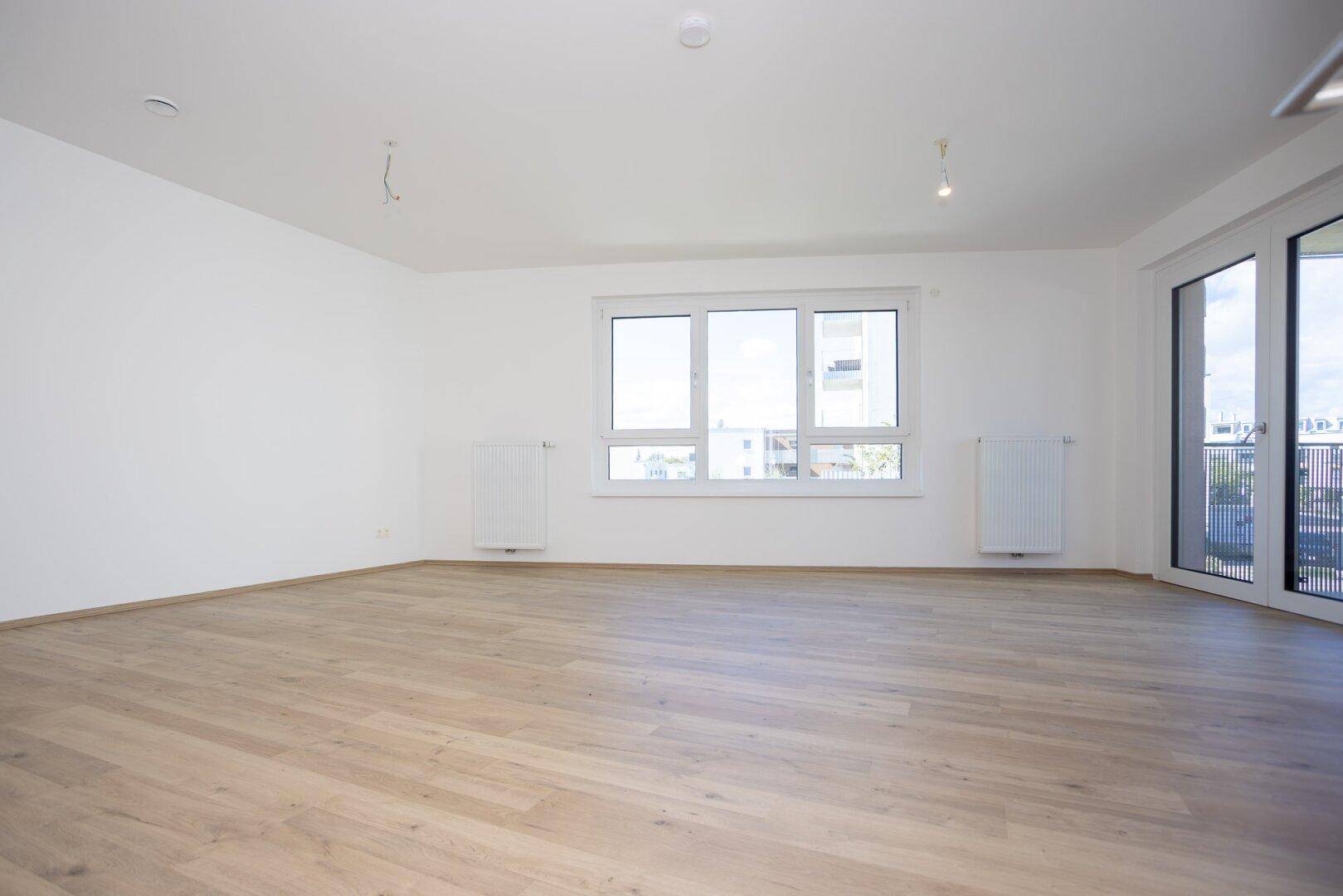 Lichtdurchflutetes Wohnzimmer mit großen Fenstern, hellem Laminatboden und weißen Wänden.