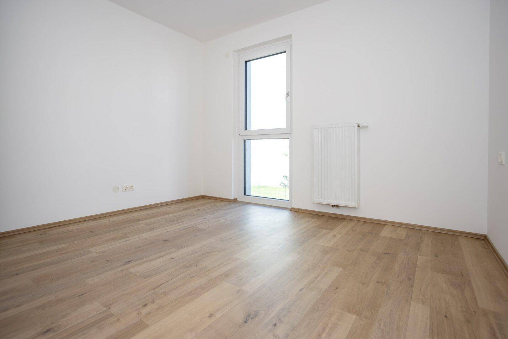 Unmöbliertes Zimmer mit hellem Laminatboden, weißen Wänden und einem hohen Fenster.