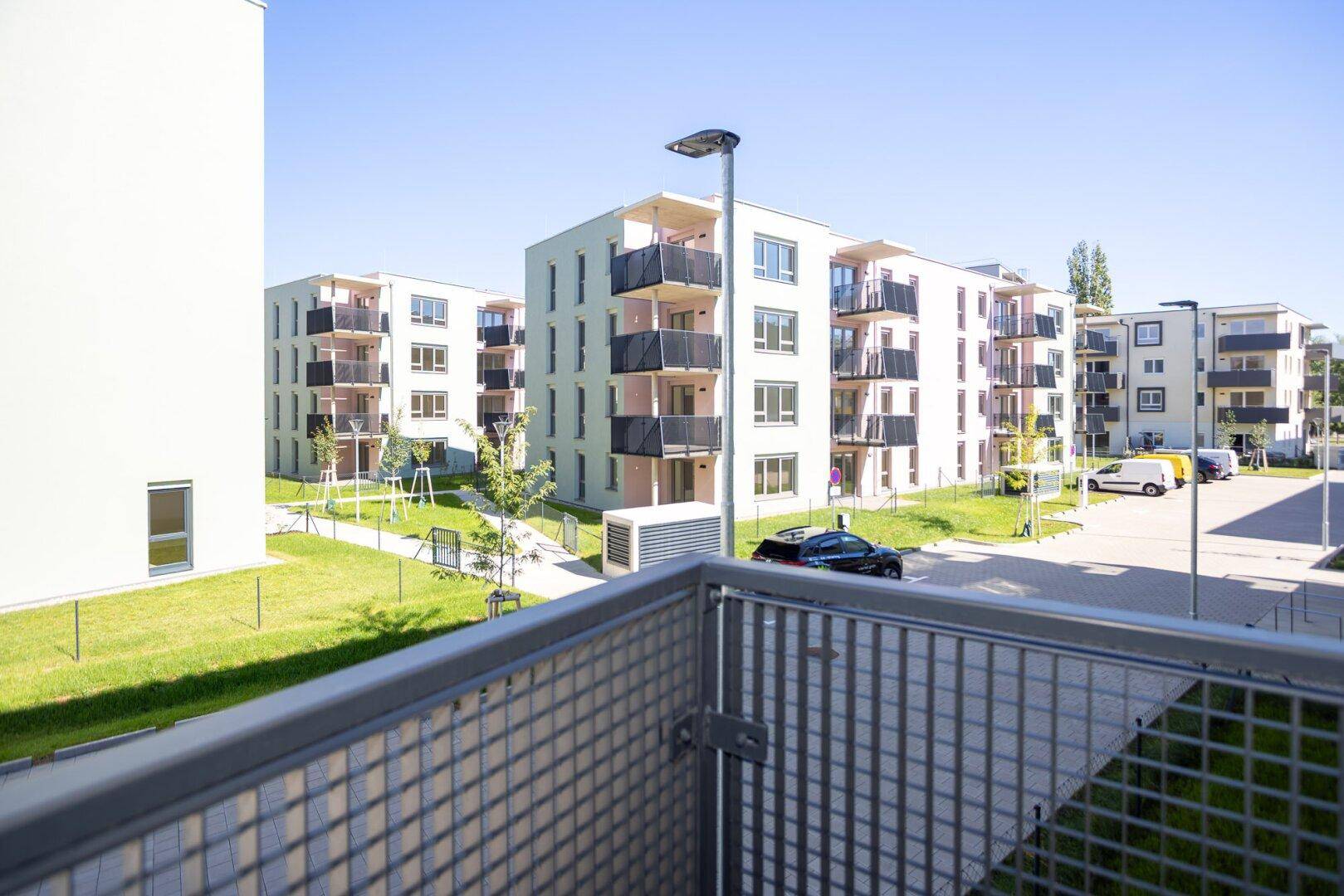 Blick vom Balkon auf moderne Wohngebäude, grüne Rasenflächen und einen Spielplatz.
