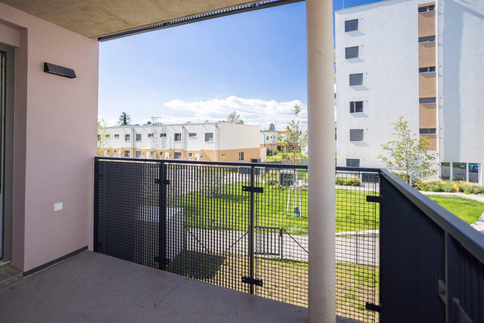 Balkon mit Blick auf eine grüne Wohnanlage und weitere Gebäude unter blauem Himmel.