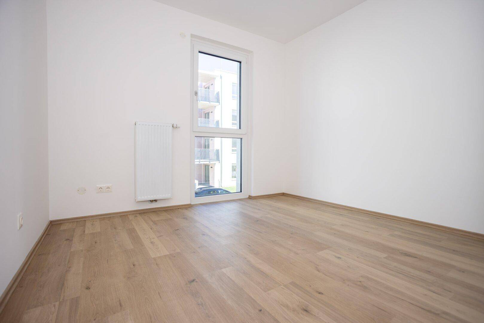 Helles Zimmer mit Laminatboden, weißen Wänden und Blick auf die umliegenden Gebäude.