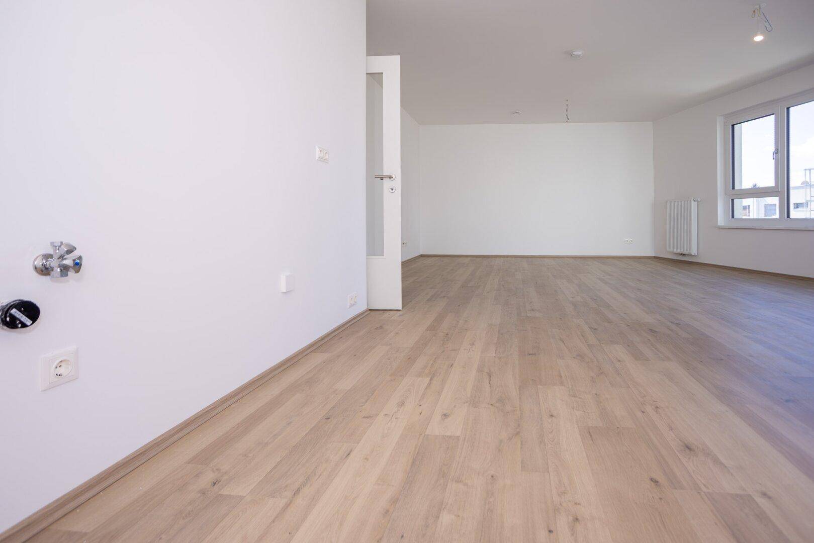Leerer Raum mit Anschlüssen für eine Küche, hellem Laminatboden und weißen Wänden.