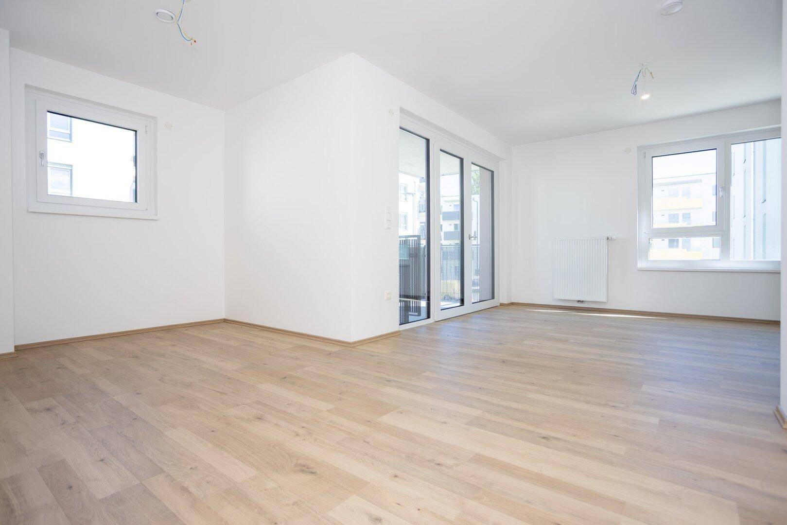 Geräumiges, helles Zimmer mit Holzboden und großen Fenstern, das viel Tageslicht hereinlässt.