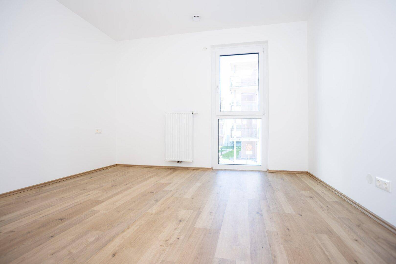 Helles Schlafzimmer mit Holzboden und einem großen Fenster, das viel Licht hereinlässt.