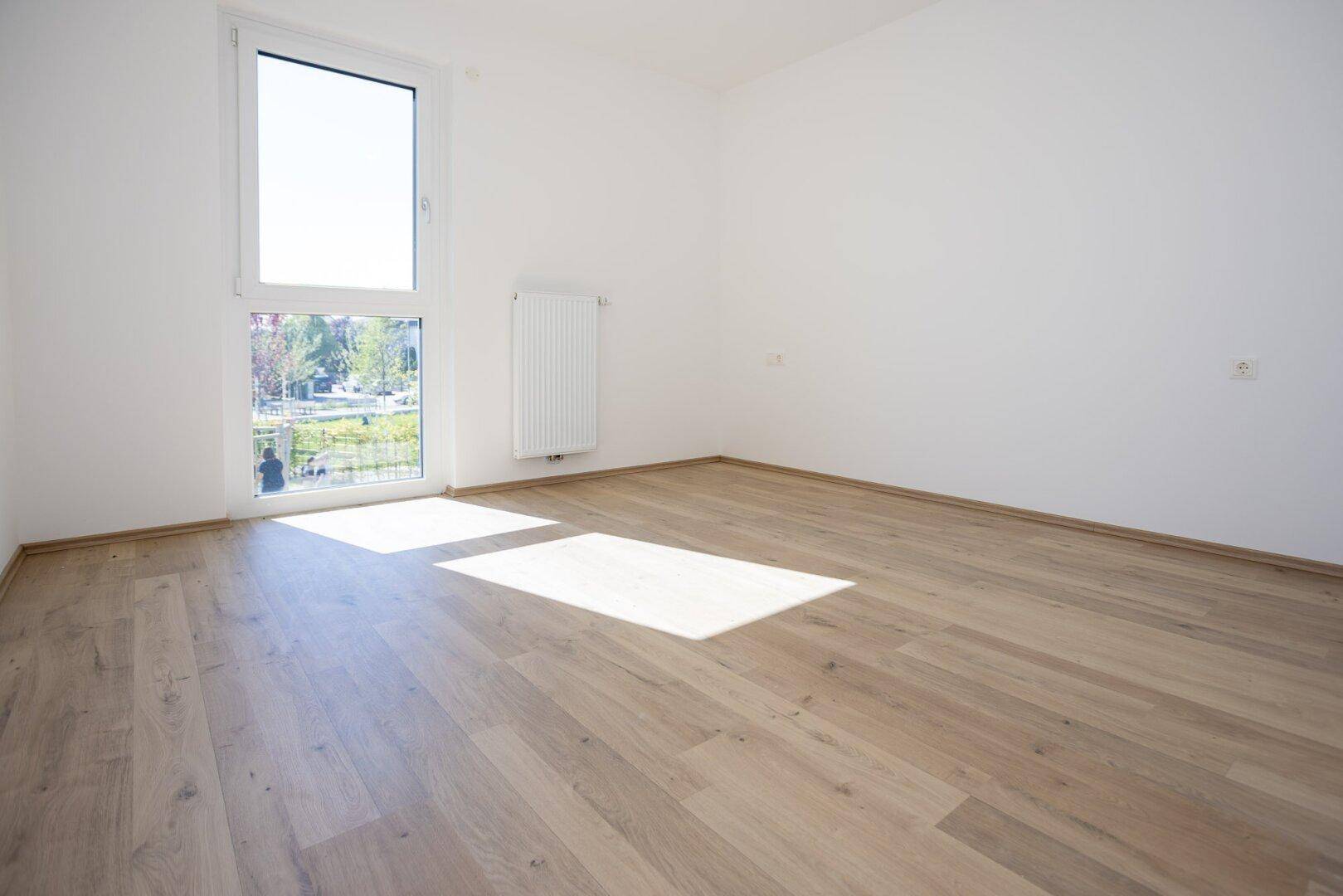 Helles Schlafzimmer mit Laminatboden und großem Fenster, das viel Licht hereinlässt.