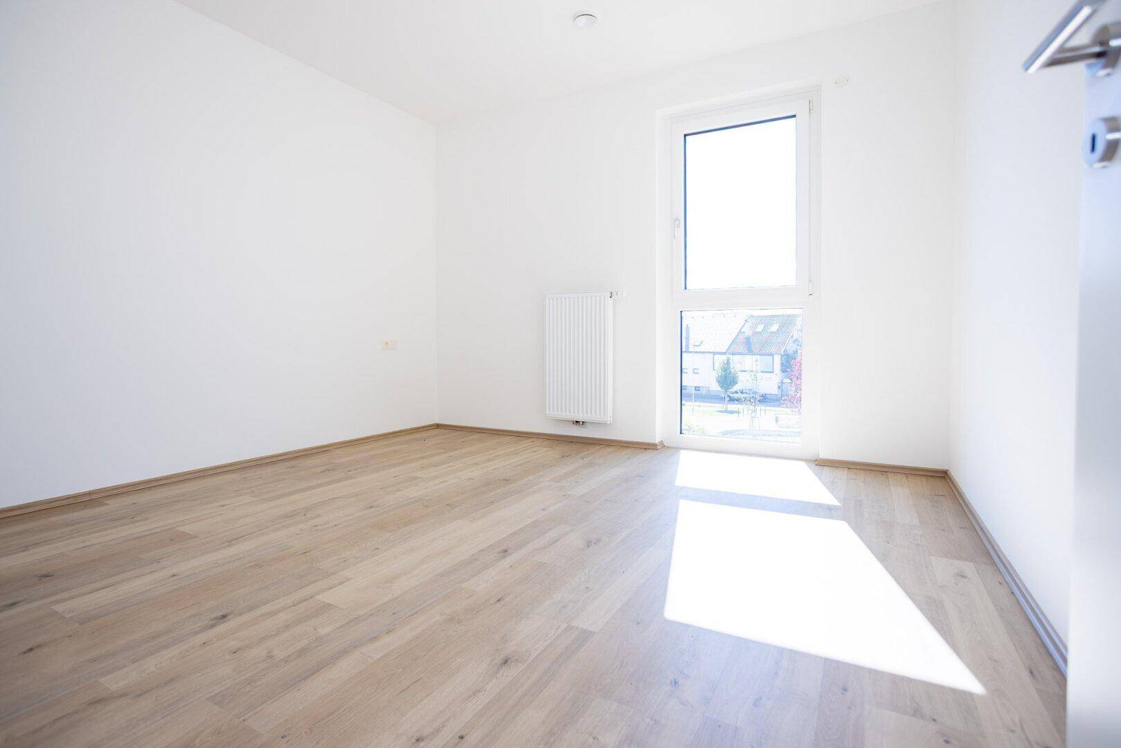 Sonniger Raum mit Holzboden und großem Fenster, ideal als Schlaf- oder Arbeitszimmer.