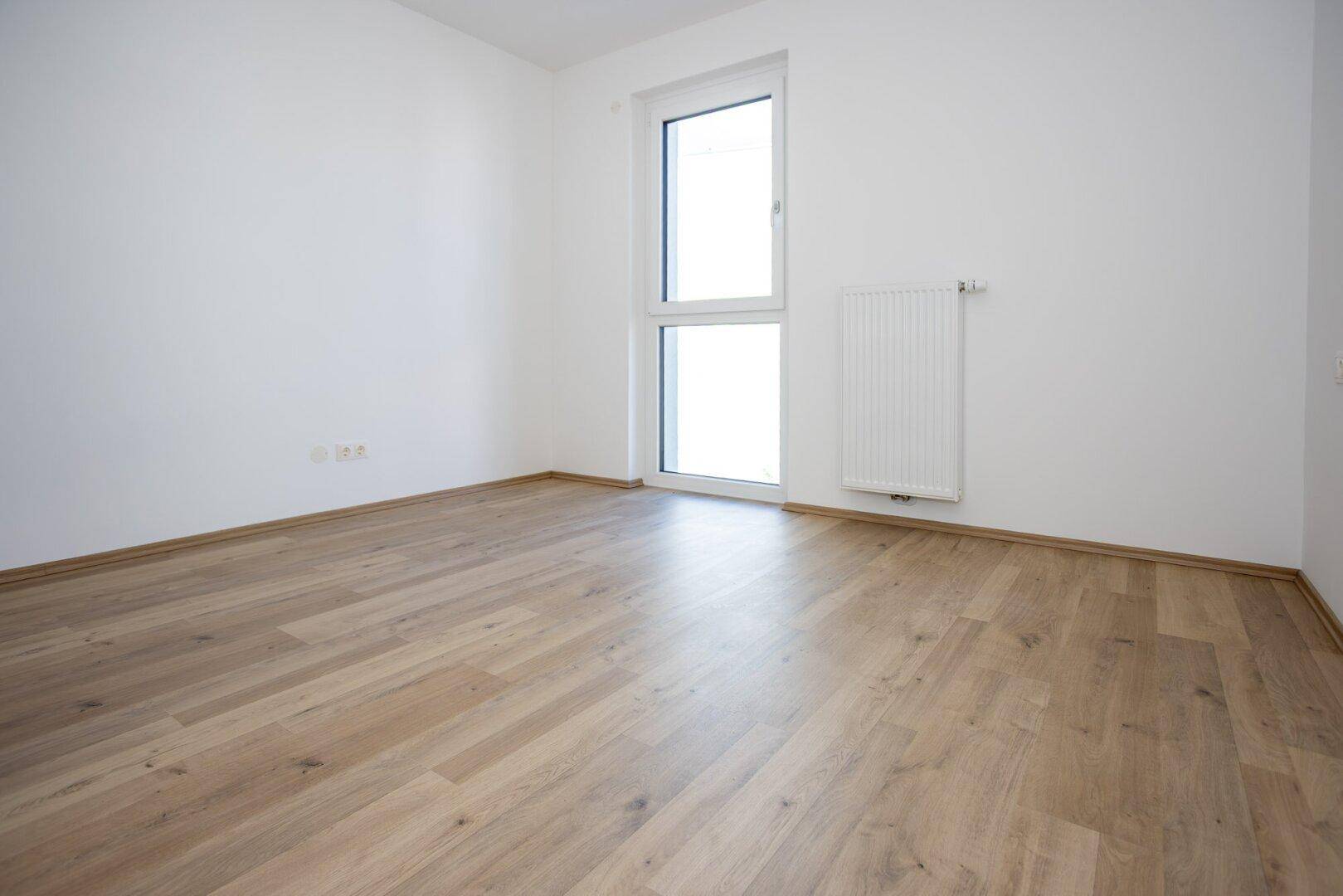 Leeres Zimmer mit hellem Laminatboden und einem hohen Fenster für viel Tageslicht.