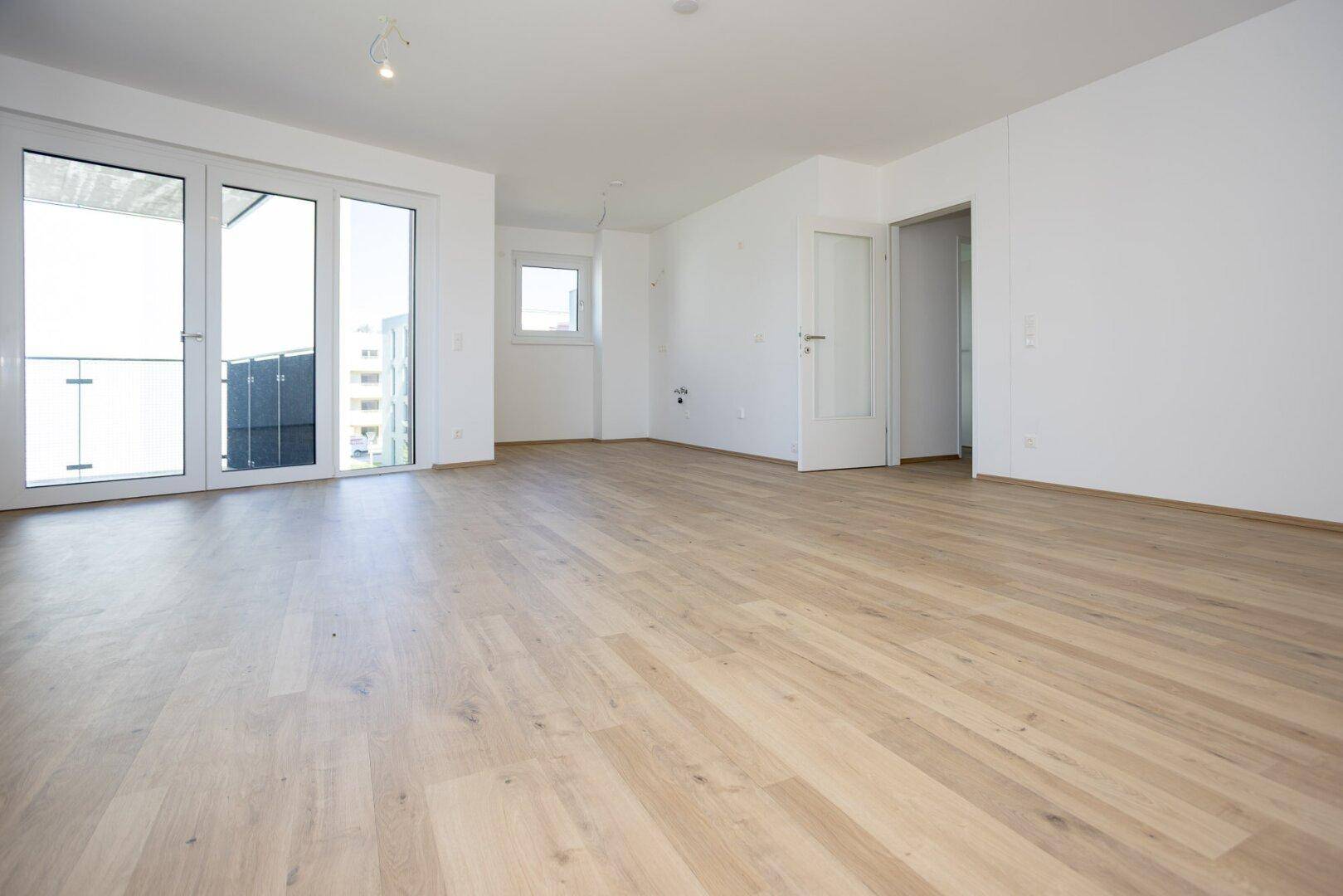 Großes, leeres Wohnzimmer mit hellem Laminatboden und Zugang zum Balkon.