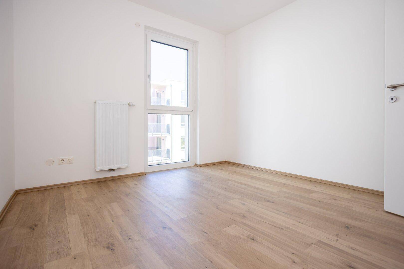 Modernes, leeres Zimmer mit hellem Laminatboden und einem hohen Fenster.