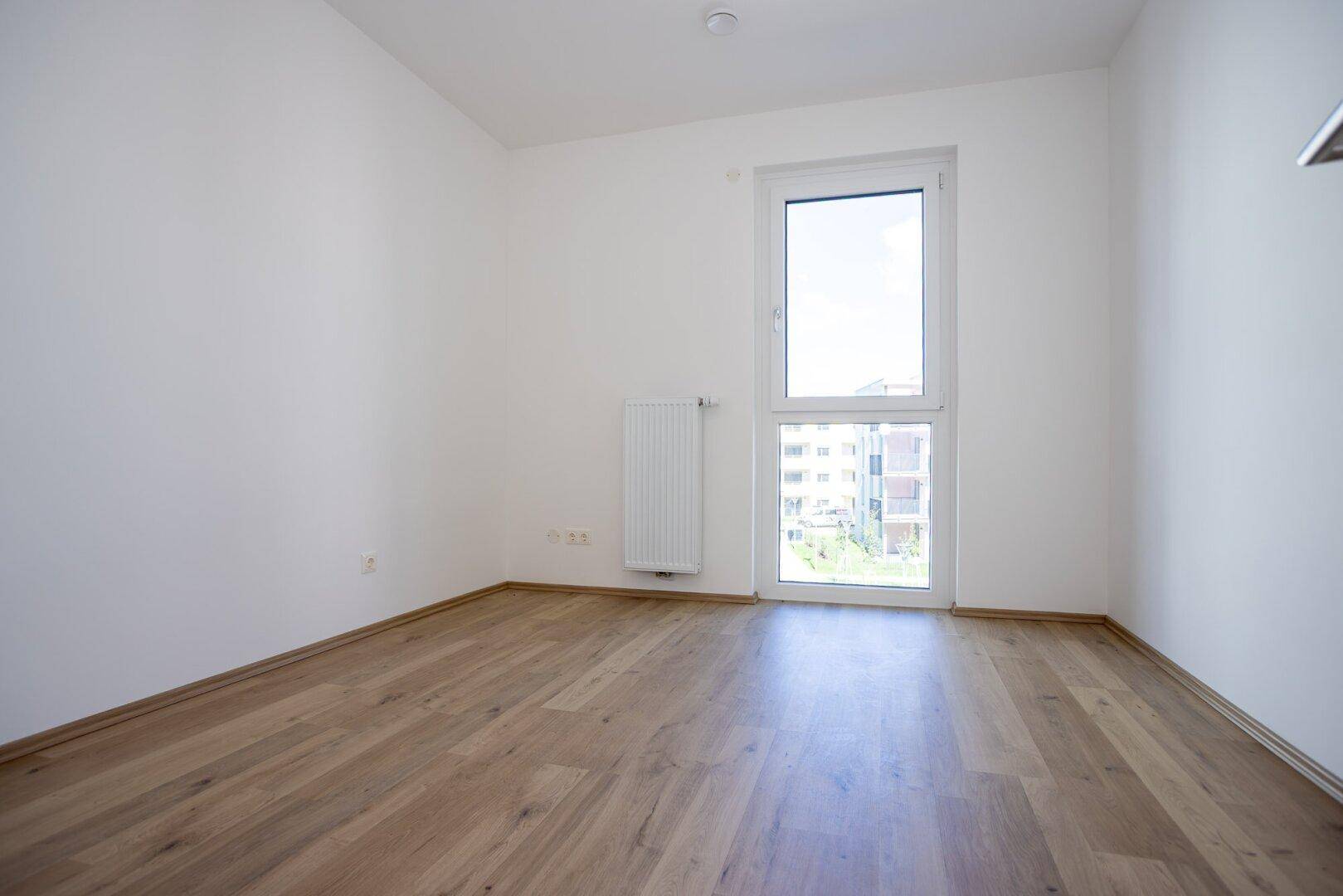 Freundliches Zimmer mit Laminatboden und einem großen Fenster mit Blick ins Grüne.