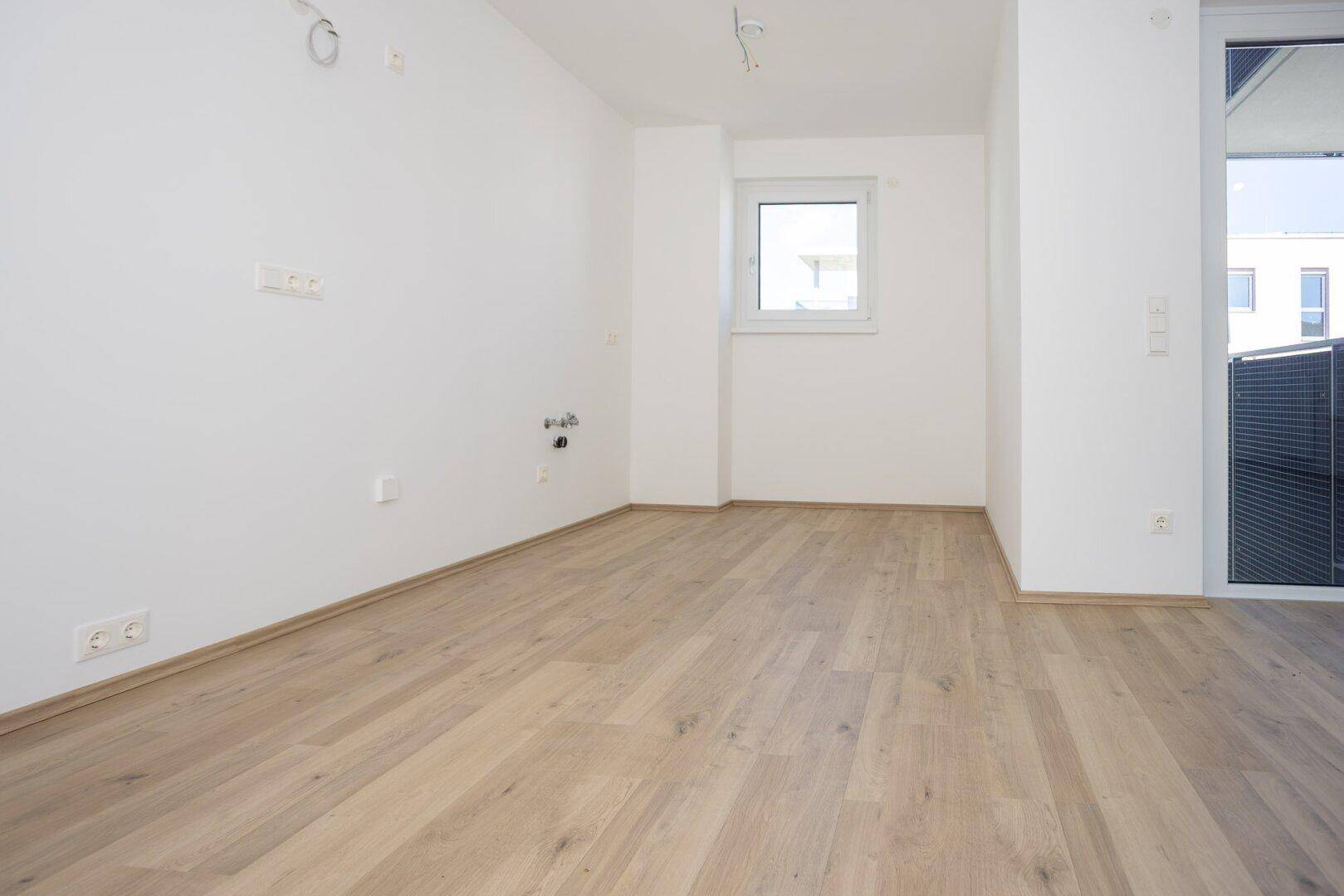 Heller Raum mit Holzboden, Fenster und Anschlüssen für eine zukünftige Küche.