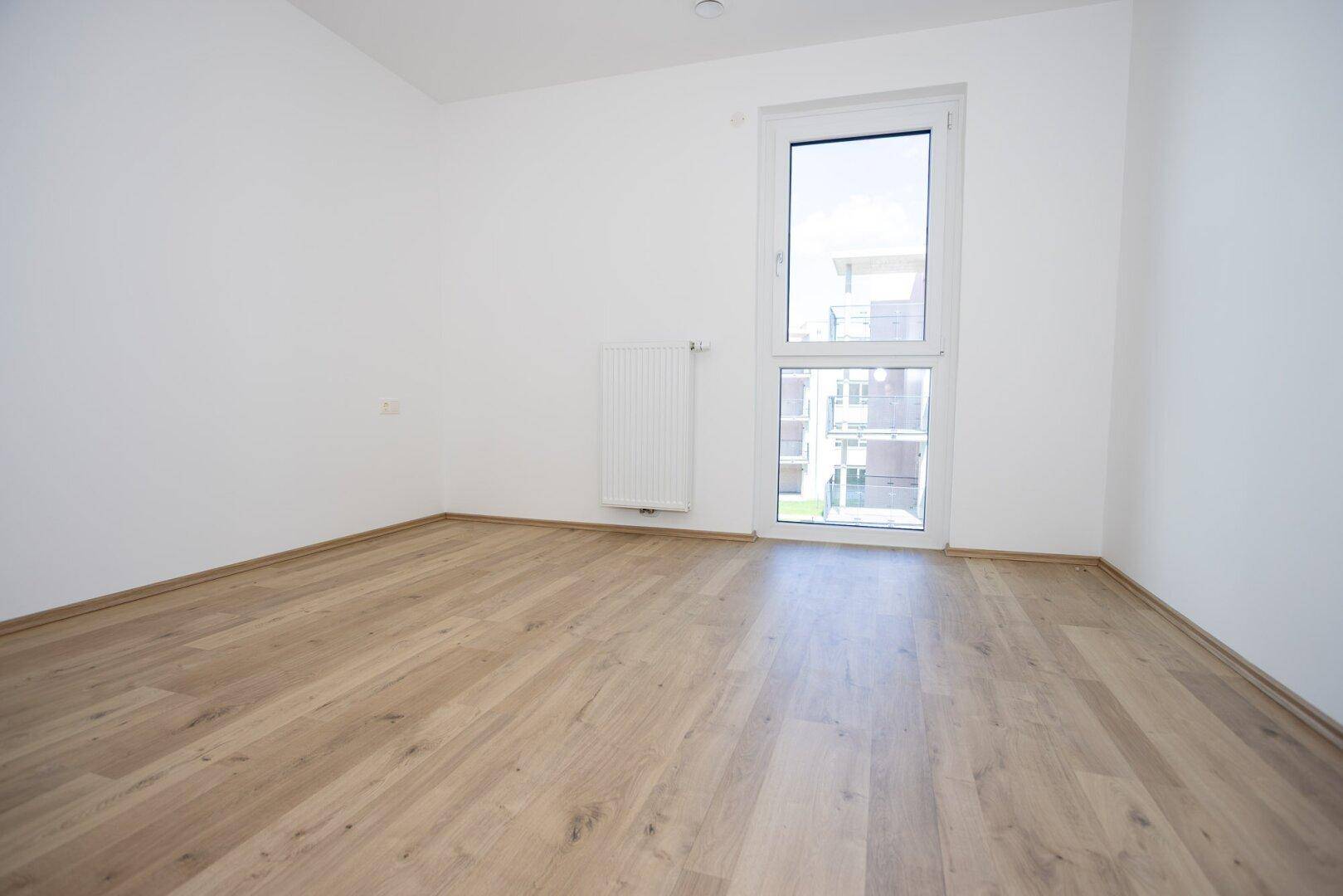 Helles Schlafzimmer mit Holzboden, großem Fenster und Heizkörper.