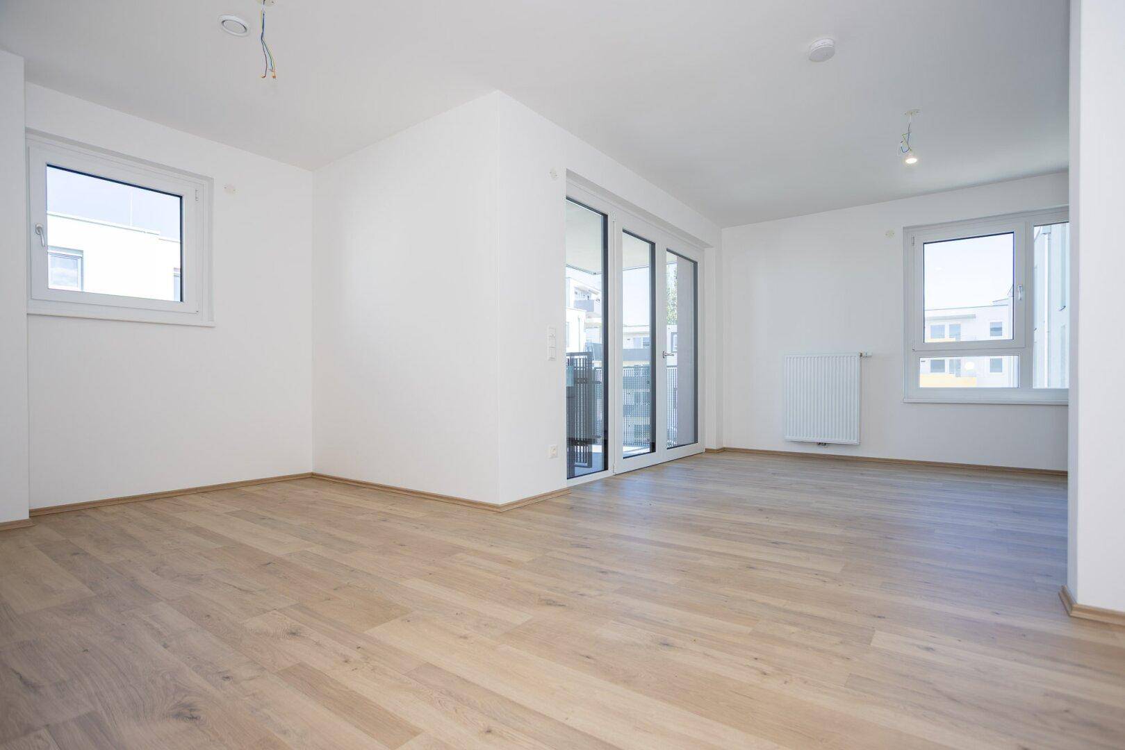 Geräumiges Wohnzimmer mit hellem Holzboden und viel Tageslicht durch große Fenster.