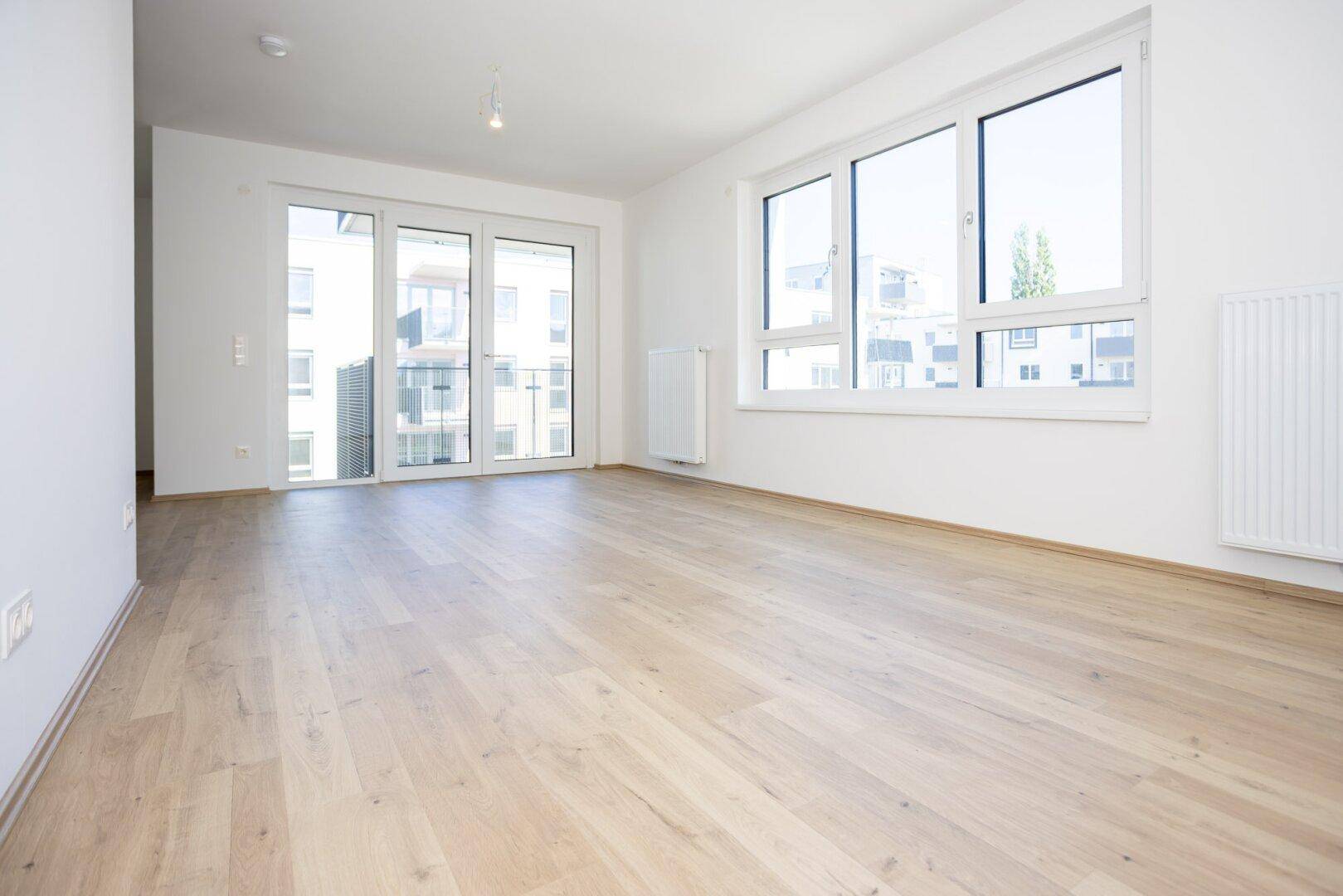 Helles Wohnzimmer mit großen Fenstern, hellem Holzboden und Zugang zum Balkon.