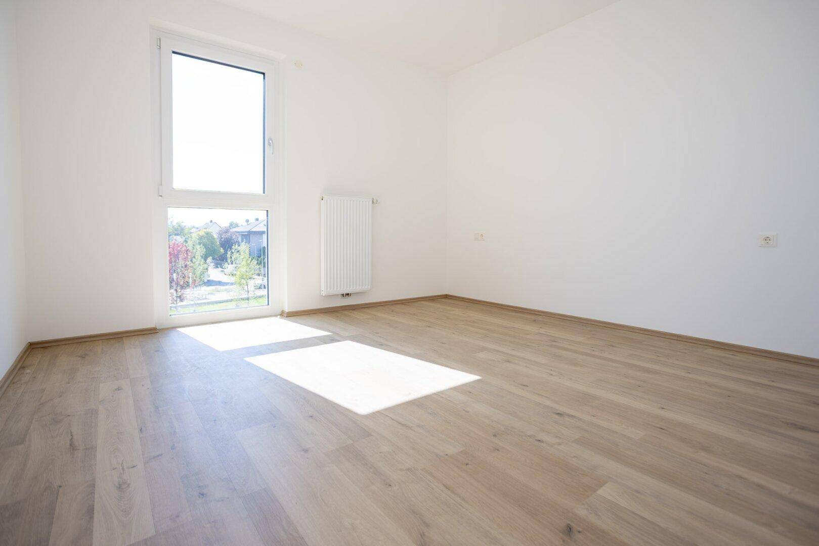 Helles Schlafzimmer mit Laminatboden und großem Fenster mit Blick ins Grüne.
