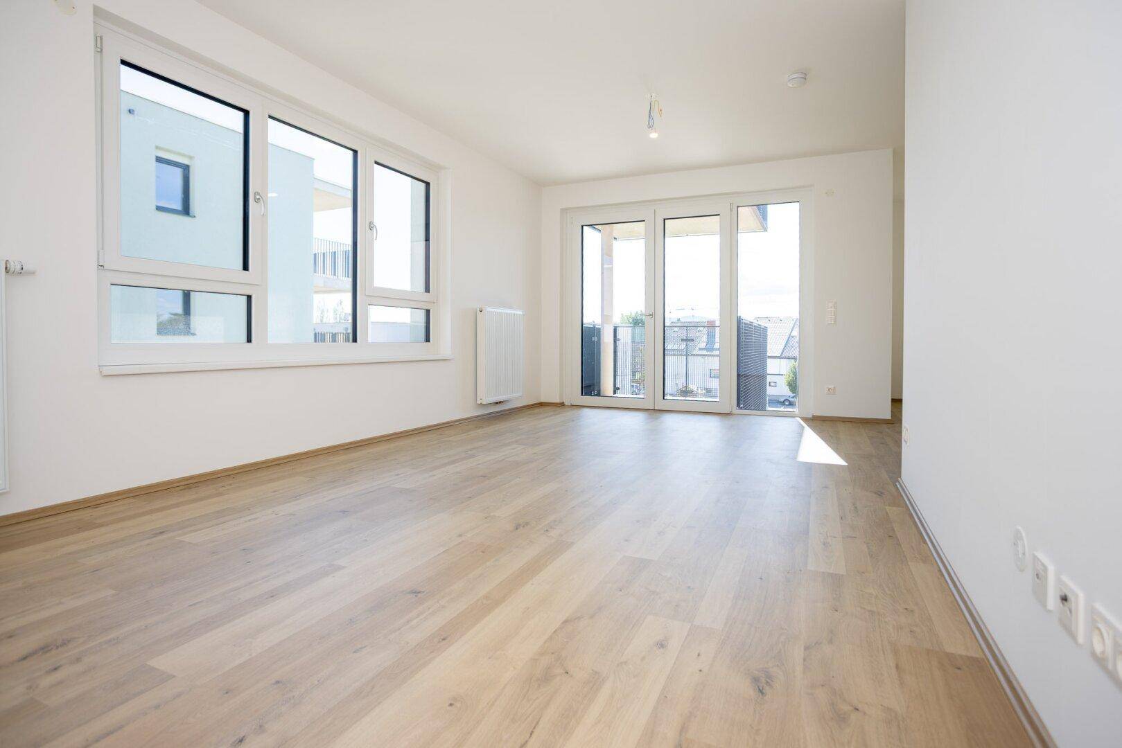 Heller Wohnraum mit modernem Laminatboden, großen Fenstern und Zugang zum Balkon.