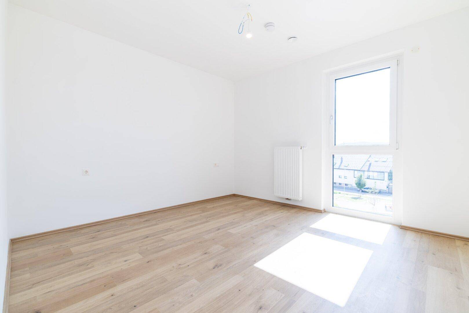 Helles Schlafzimmer mit Laminatboden und großem Fenster, das viel Tageslicht hereinlässt.