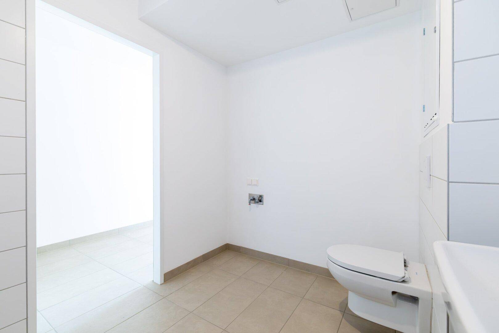Heller Toilettenbereich mit Wand-WC und Fliesenboden, angrenzend an einen weiteren Raum.