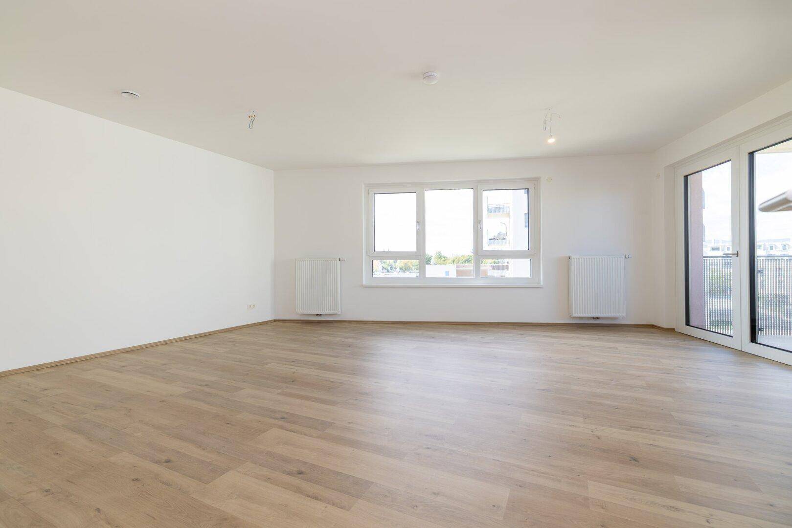 Geräumiges, leeres Wohnzimmer mit großen Fenstern und hellem Laminatboden.