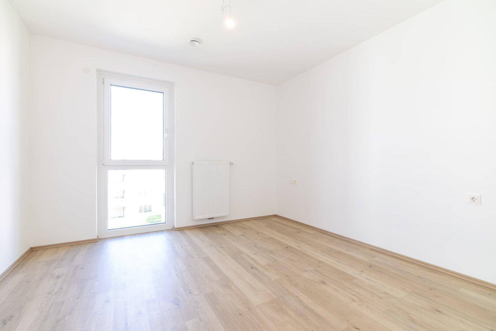 Helles, leeres Schlafzimmer mit Laminatboden und großem Fenster, bereit zur Einrichtung.