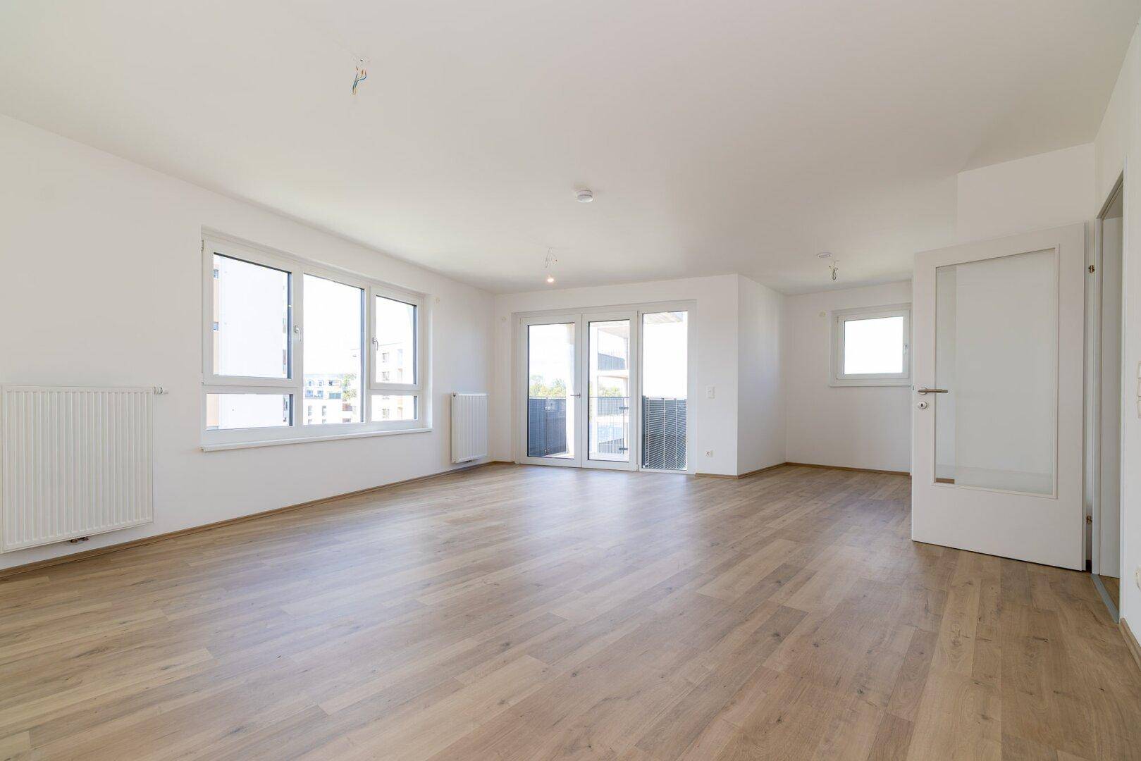 Lichtdurchflutetes Wohnzimmer mit hellem Laminatboden, großen Fenstern und Balkonzugang.