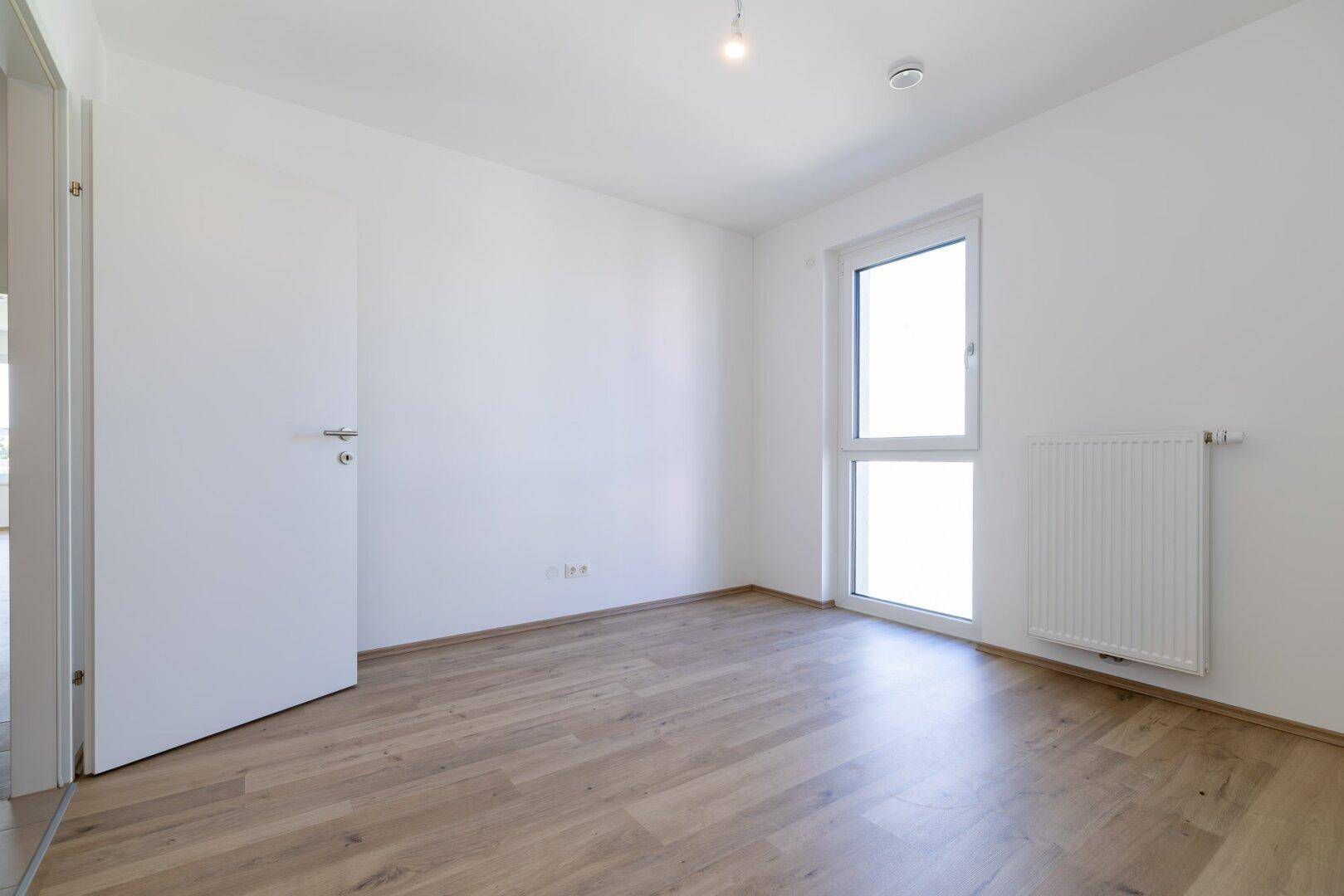 Geräumiges, leeres Schlafzimmer mit hellem Laminatboden und Fenster, ideal für viel Tageslicht.