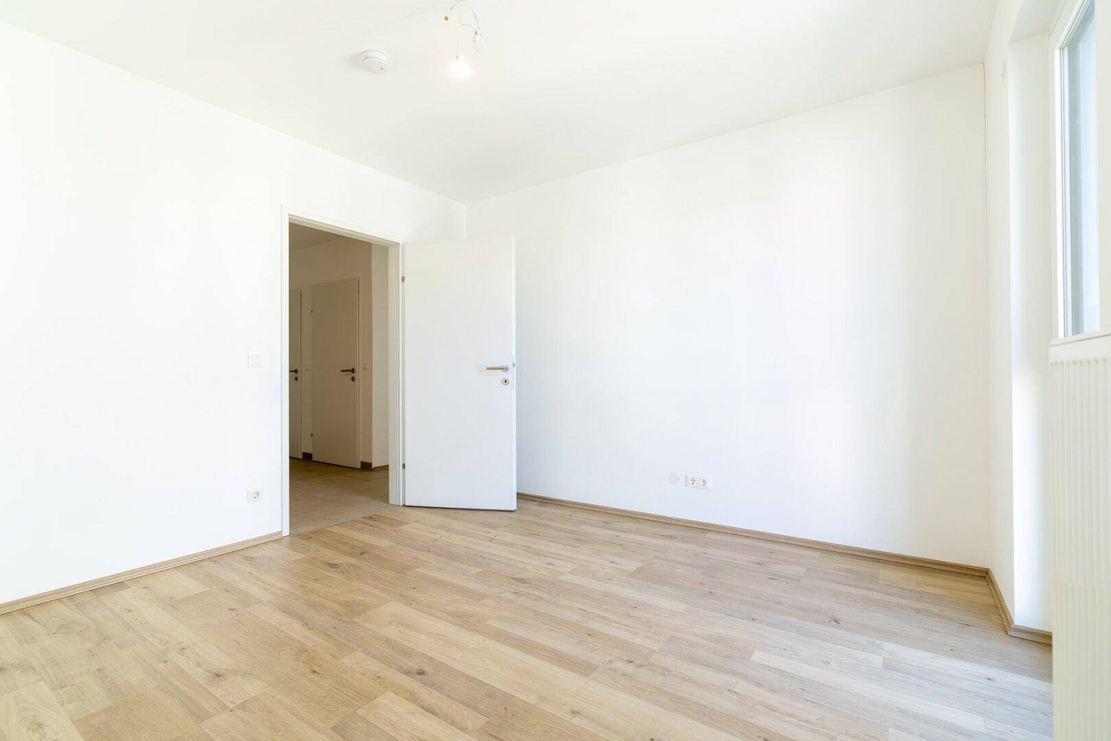 Helles, leeres Schlafzimmer mit Laminatboden und offener Tür zu weiteren Räumen.