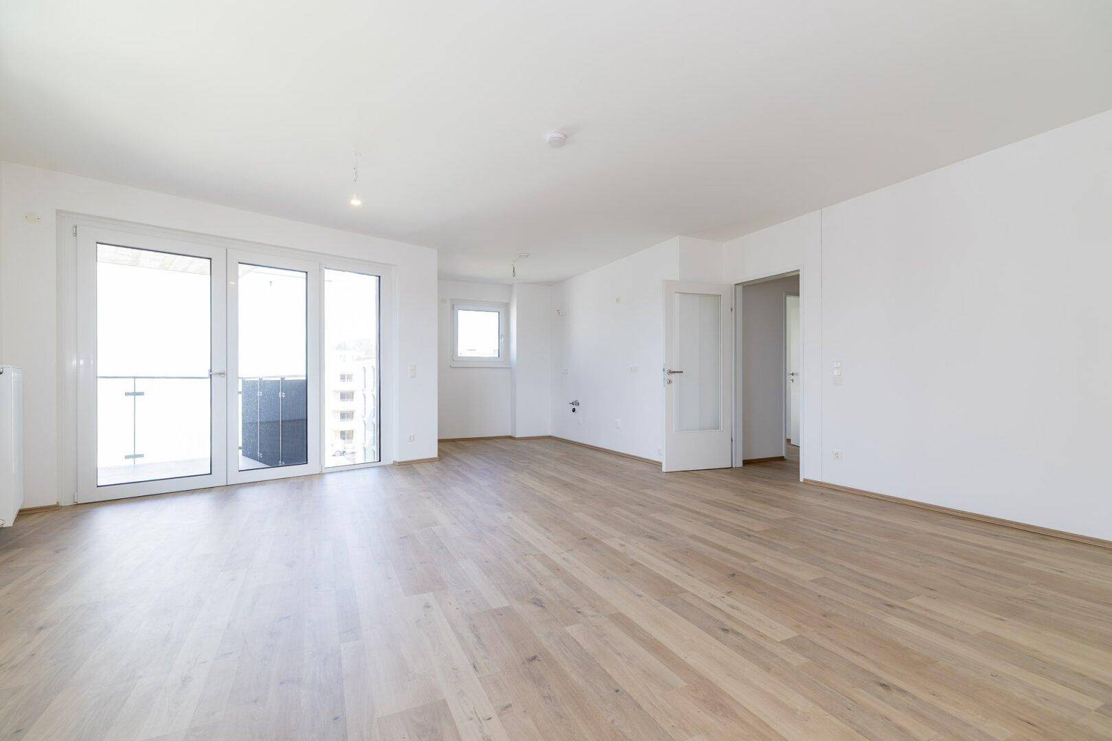 Helles, leeres Wohnzimmer mit hellem Laminatboden und Zugang zu einem Balkon.