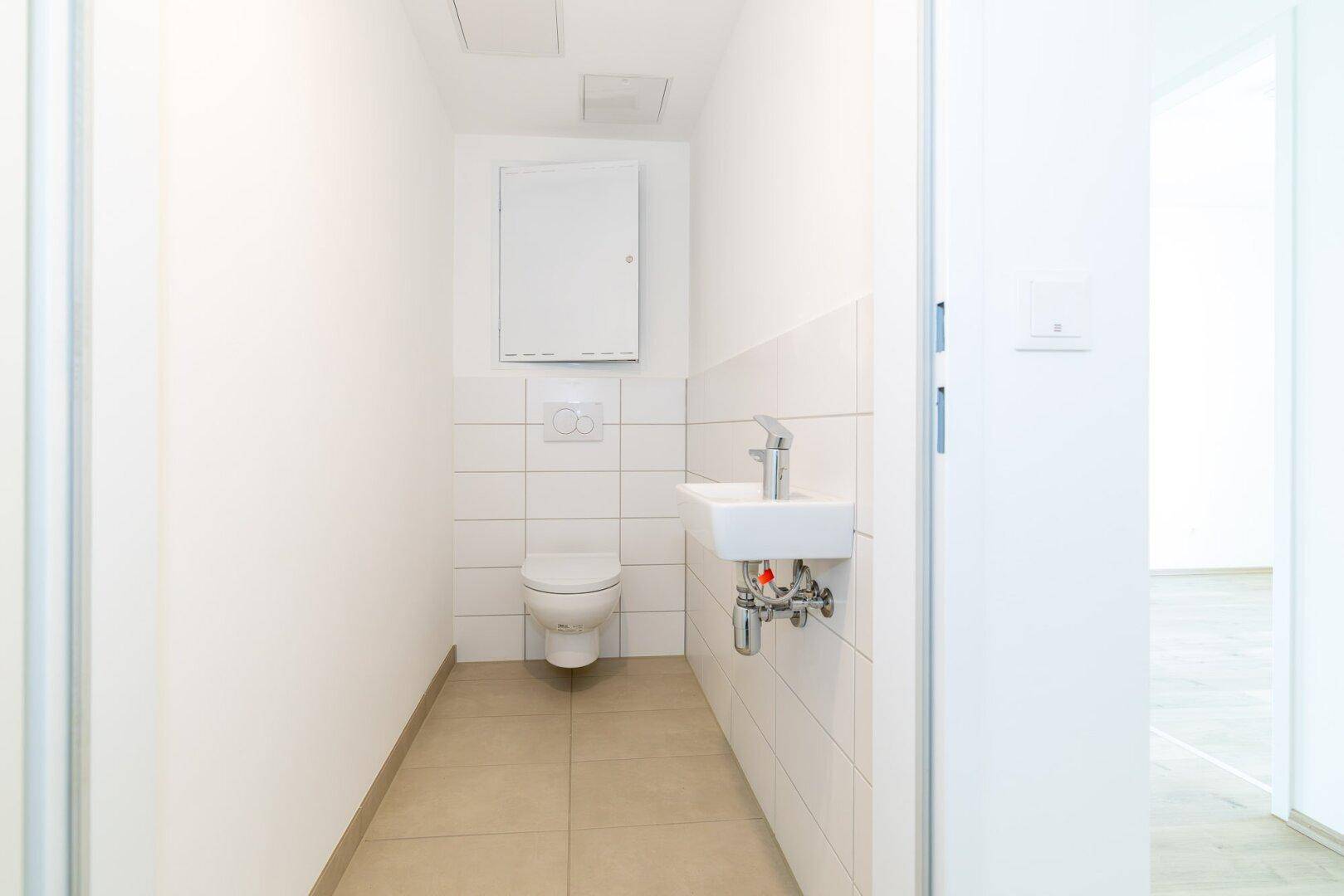 Helles Gäste-WC mit Wand-WC, kleinem Waschbecken und beigefarbenen Bodenfliesen.