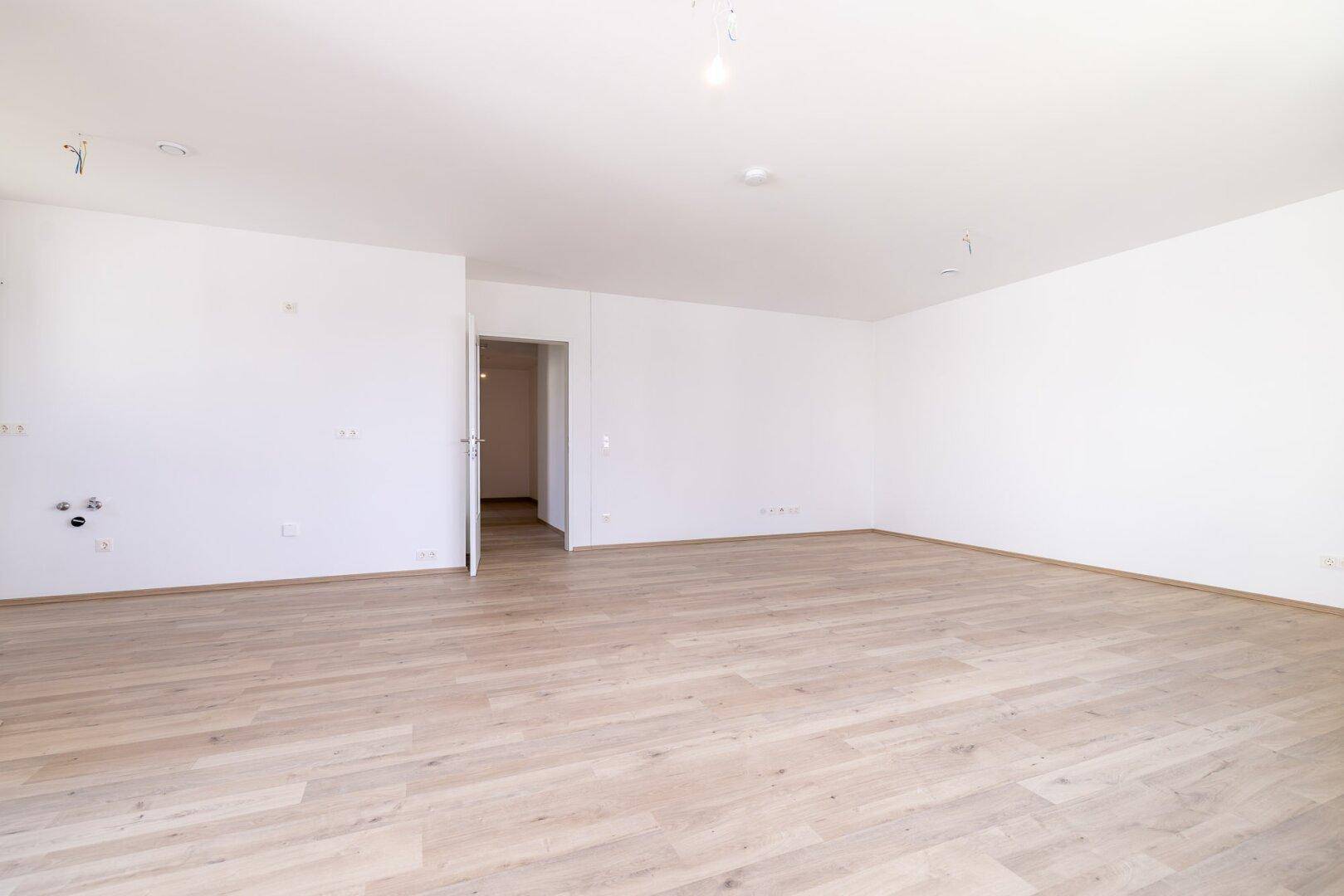 Geräumiges, leeres Wohnzimmer mit hellem Laminatboden und offenem Zugang zu weiteren Räumen.