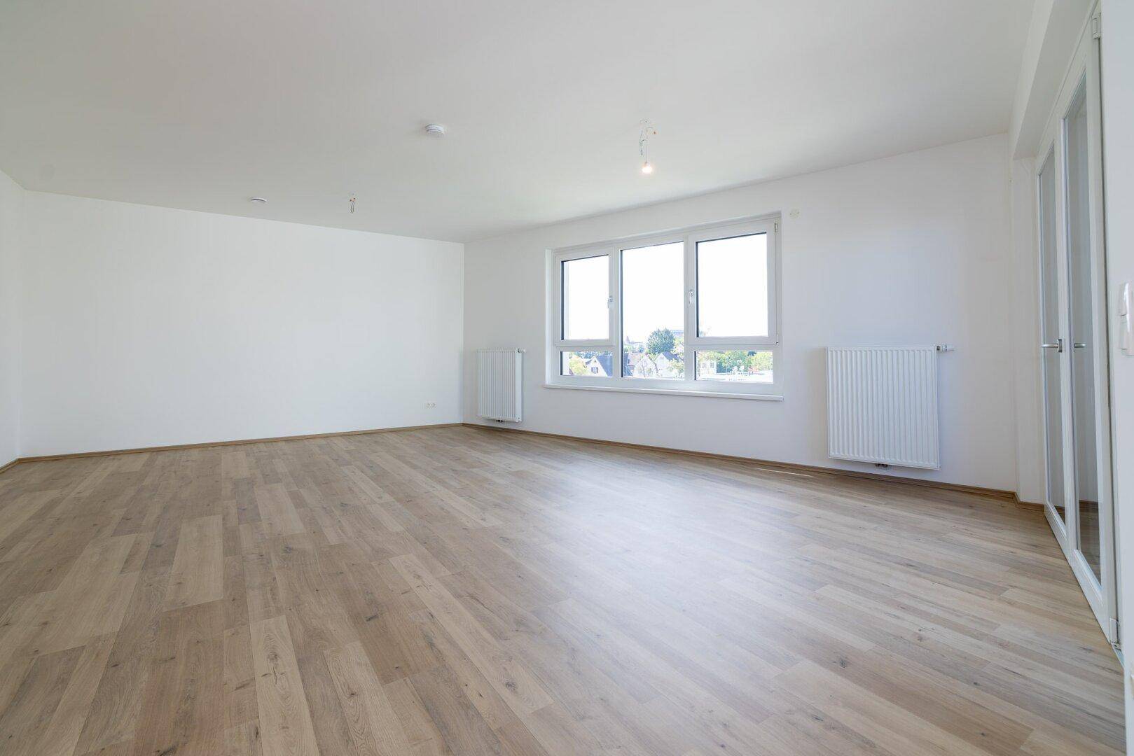 Helles, leeres Wohnzimmer mit Laminatboden und großen Fenstern, bereit zur individuellen Gestaltung.