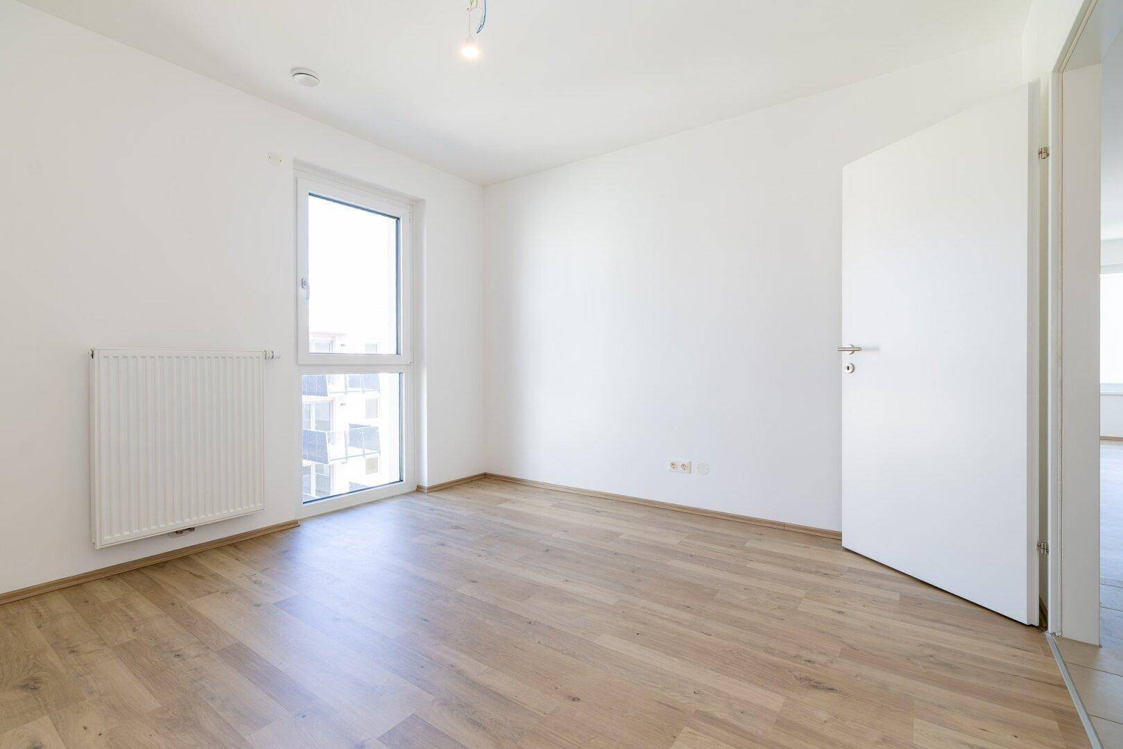 Geräumiges Schlafzimmer mit hellem Laminatboden, weißen Wänden und viel Tageslicht.