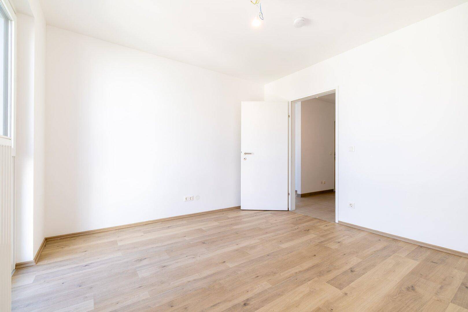 Leeres Schlafzimmer mit Laminatboden, weißen Wänden und offener Tür zu einem weiteren Raum.