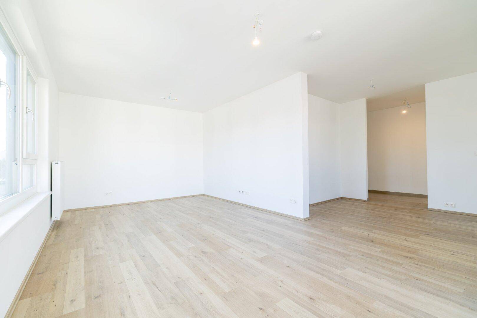 Offener Wohnbereich mit hellem Laminatboden, weißen Wänden und viel natürlichem Licht.