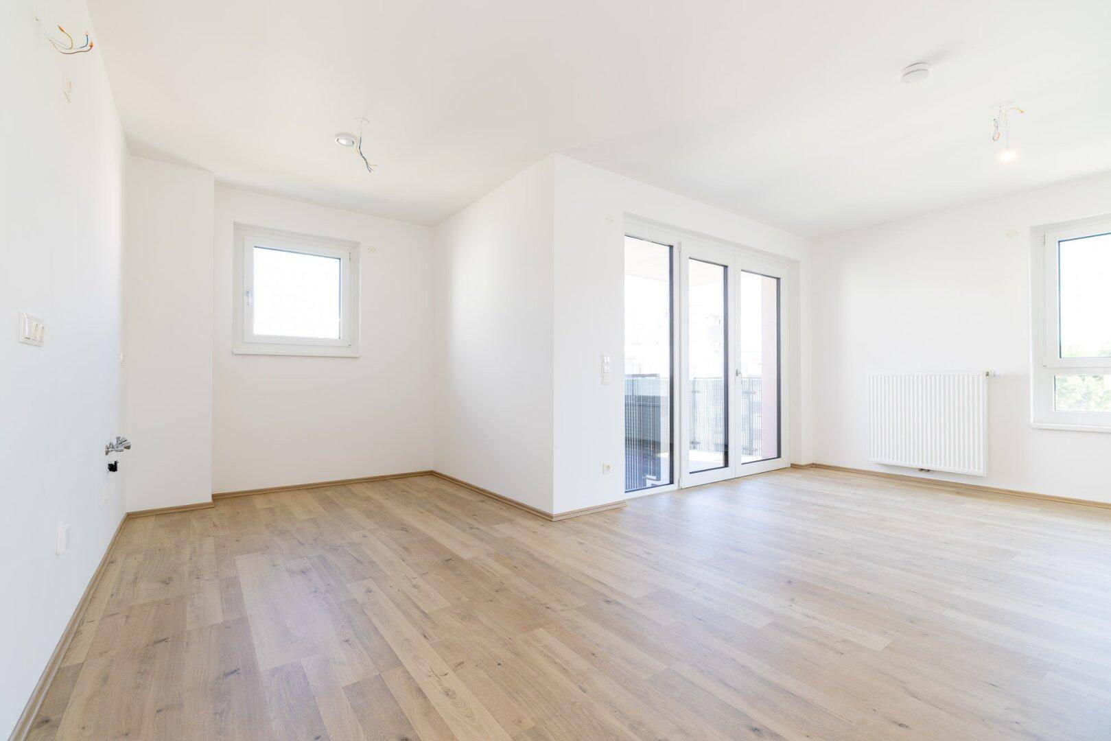 Lichtdurchflutetes Wohnzimmer mit hellem Laminatboden, weißen Wänden und Balkonzugang.