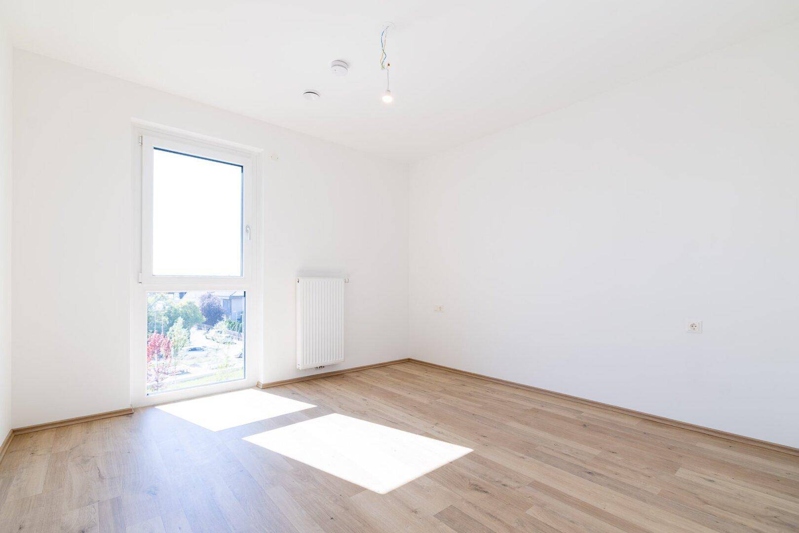 Helles Schlafzimmer mit Laminatboden und einem großen Fenster, das viel Sonnenlicht hereinlässt.