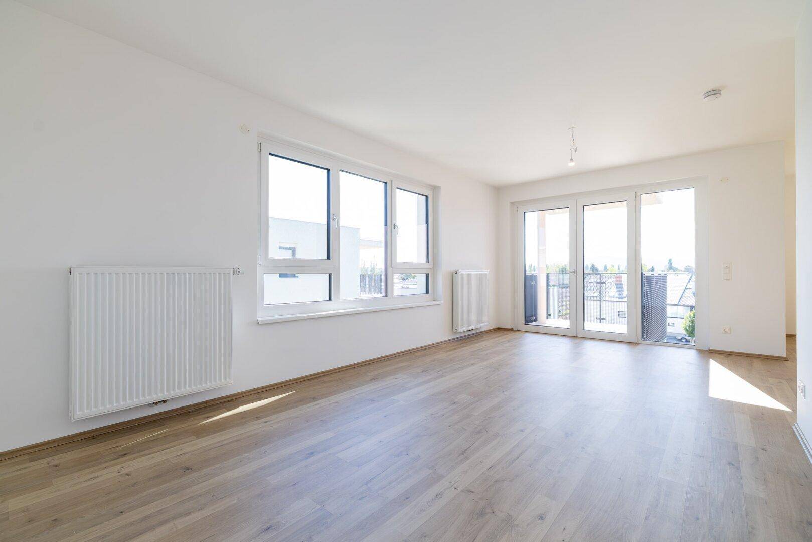 Helles, leeres Zimmer mit Laminatboden, großen Fenstern und Zugang zu einem Balkon.