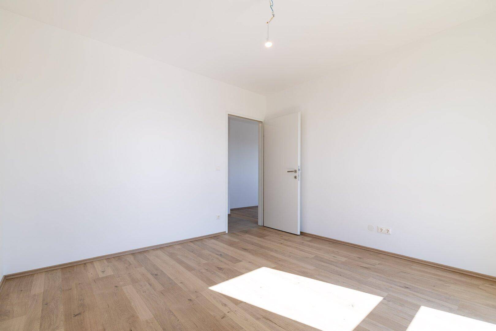 Leeres Zimmer mit Laminatboden und weiß gestrichenen Wänden, mit offener Tür zu einem Nebenraum.