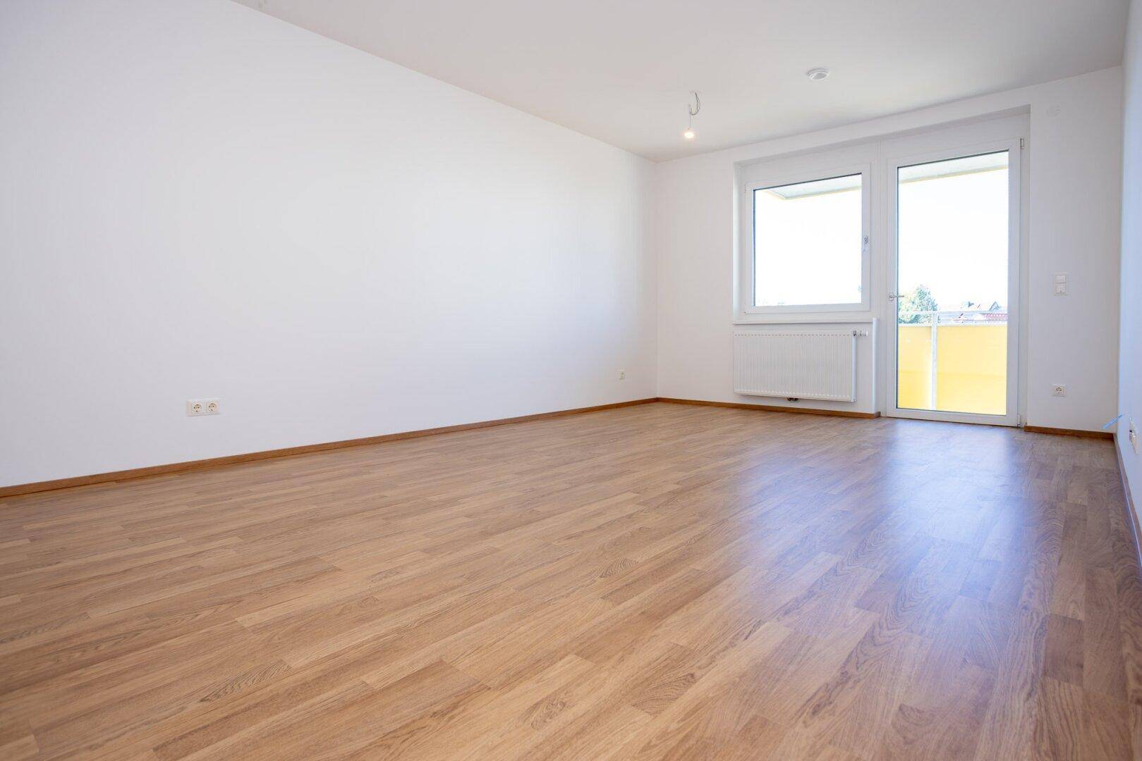 Helles, leeres Wohnzimmer mit modernem Laminatboden und Zugang zu einem Balkon.