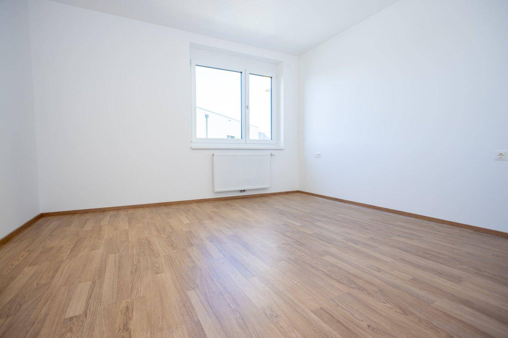 Helles Schlafzimmer mit Laminatboden, weißen Wänden und einem großen Fenster für viel Tageslicht.
