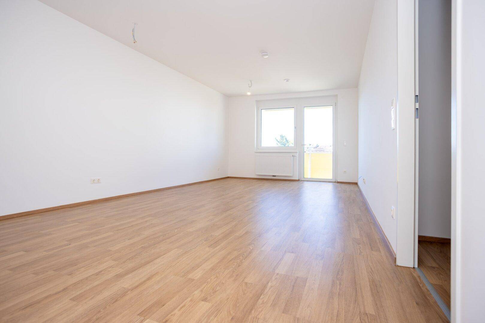 Geräumiges, unmöbliertes Wohnzimmer mit hellem Laminatboden und großen Fenstern für viel Tageslicht.