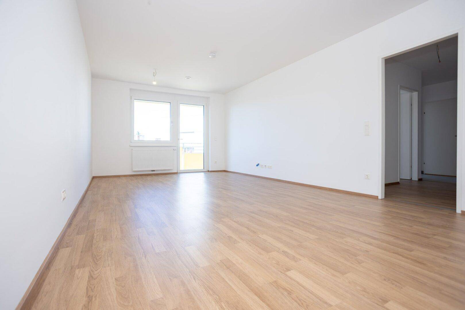 Lichtdurchflutetes Wohnzimmer mit Laminatboden, weißen Wänden und offenem Zugang zu weiteren Räumen.