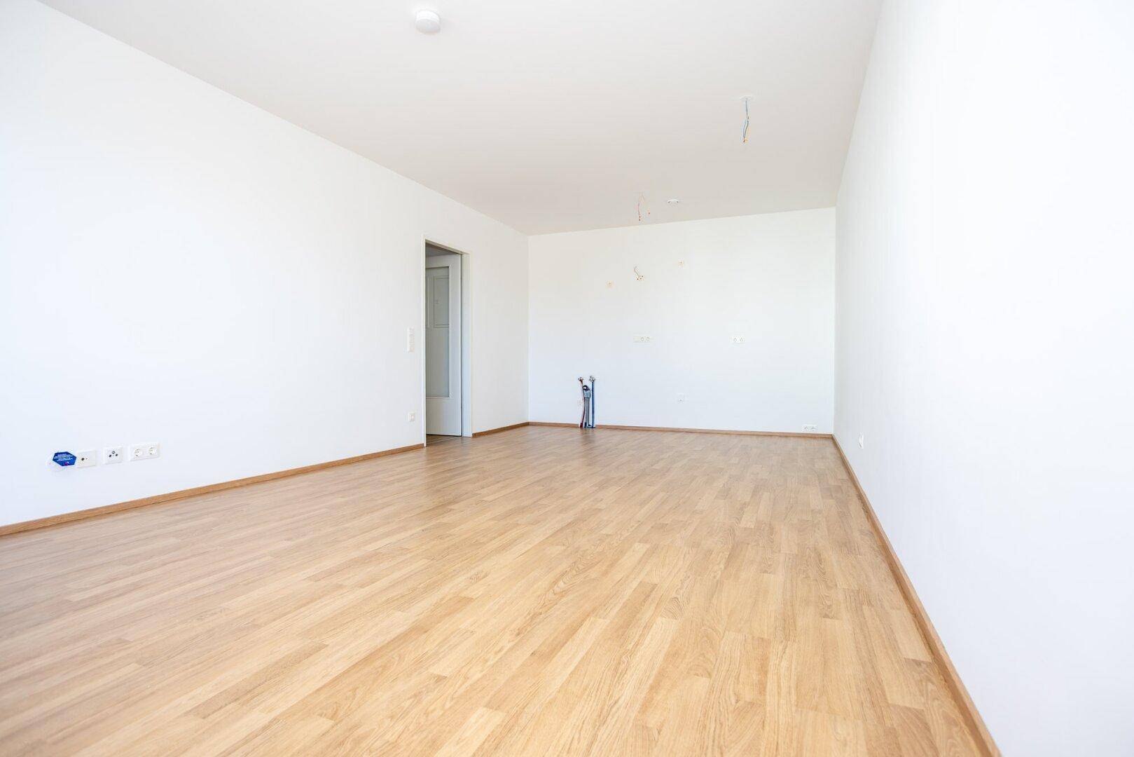 Leerer Raum mit hellem Laminatboden und weißen Wänden, vorbereitet für die Installation einer Küche.