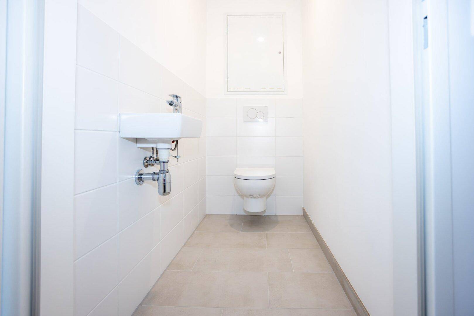 Separates Gäste-WC mit weißen Fliesen, wandhängendem WC und kleinem Handwaschbecken.