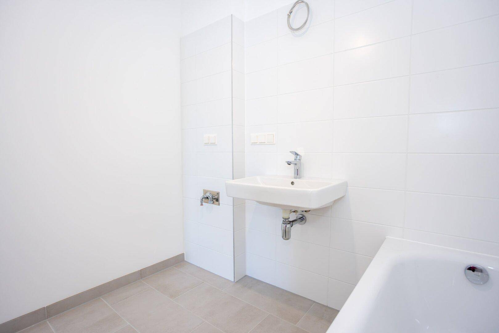 Helles Badezimmer mit weißen Fliesen, Waschbecken und Badewannenbereich, in gepflegtem Zustand.
