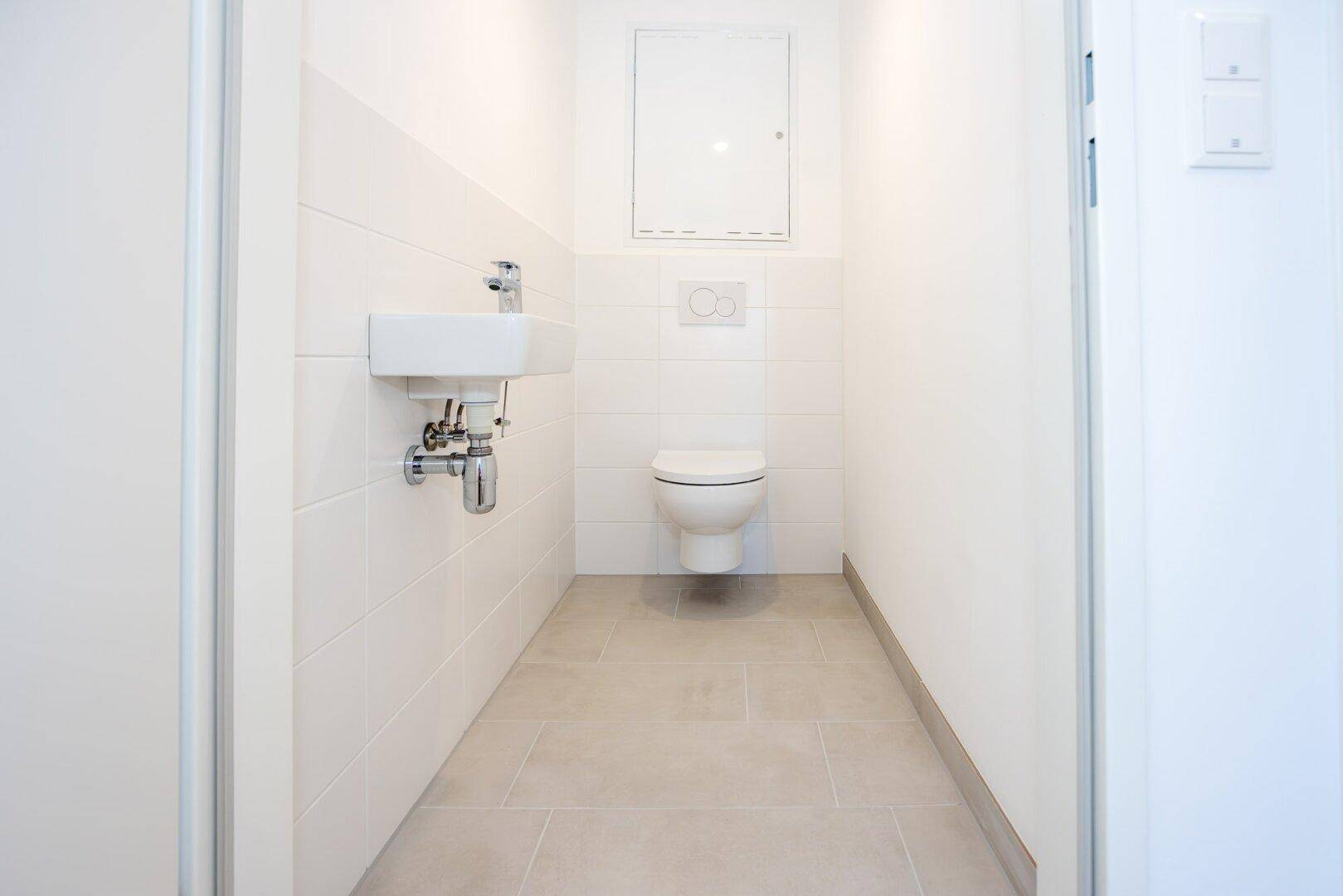 Separates Gäste-WC mit wandhängender Toilette und kleinem Waschbecken, hell gefliest.