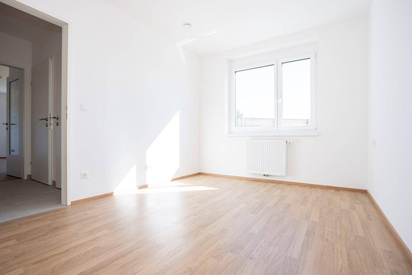 Freundliches Zimmer mit Laminatboden und großem Fenster, das den Raum mit Sonnenlicht füllt.
