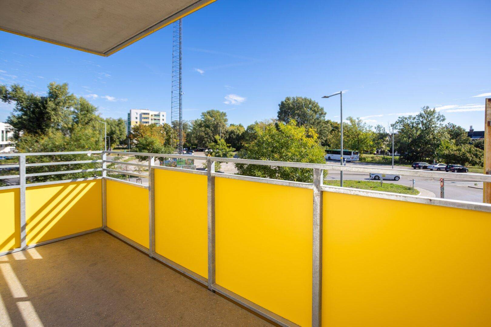 Geräumiger Balkon mit leuchtend gelben Paneelen und weitem Blick auf die Straße und Bäume.