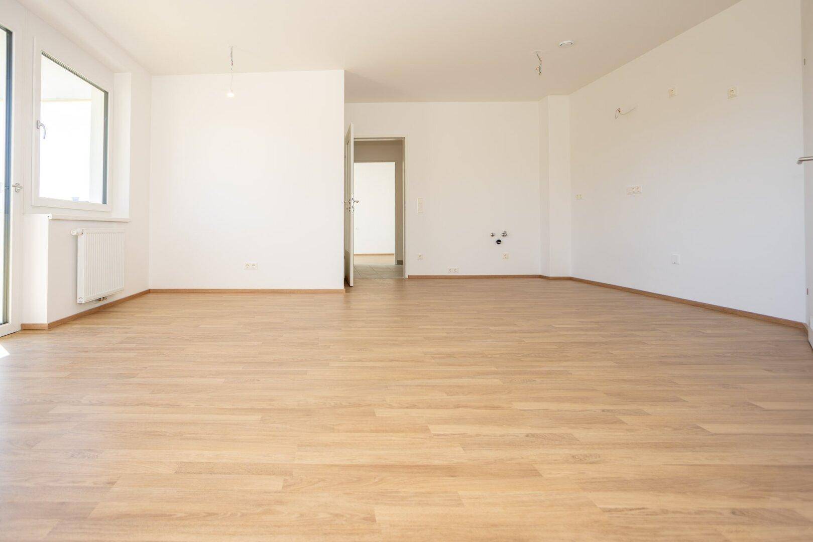 Geräumiges, leeres Zimmer mit hellem Laminatboden und weiß gestrichenen Wänden, bereit zur Einrichtung.