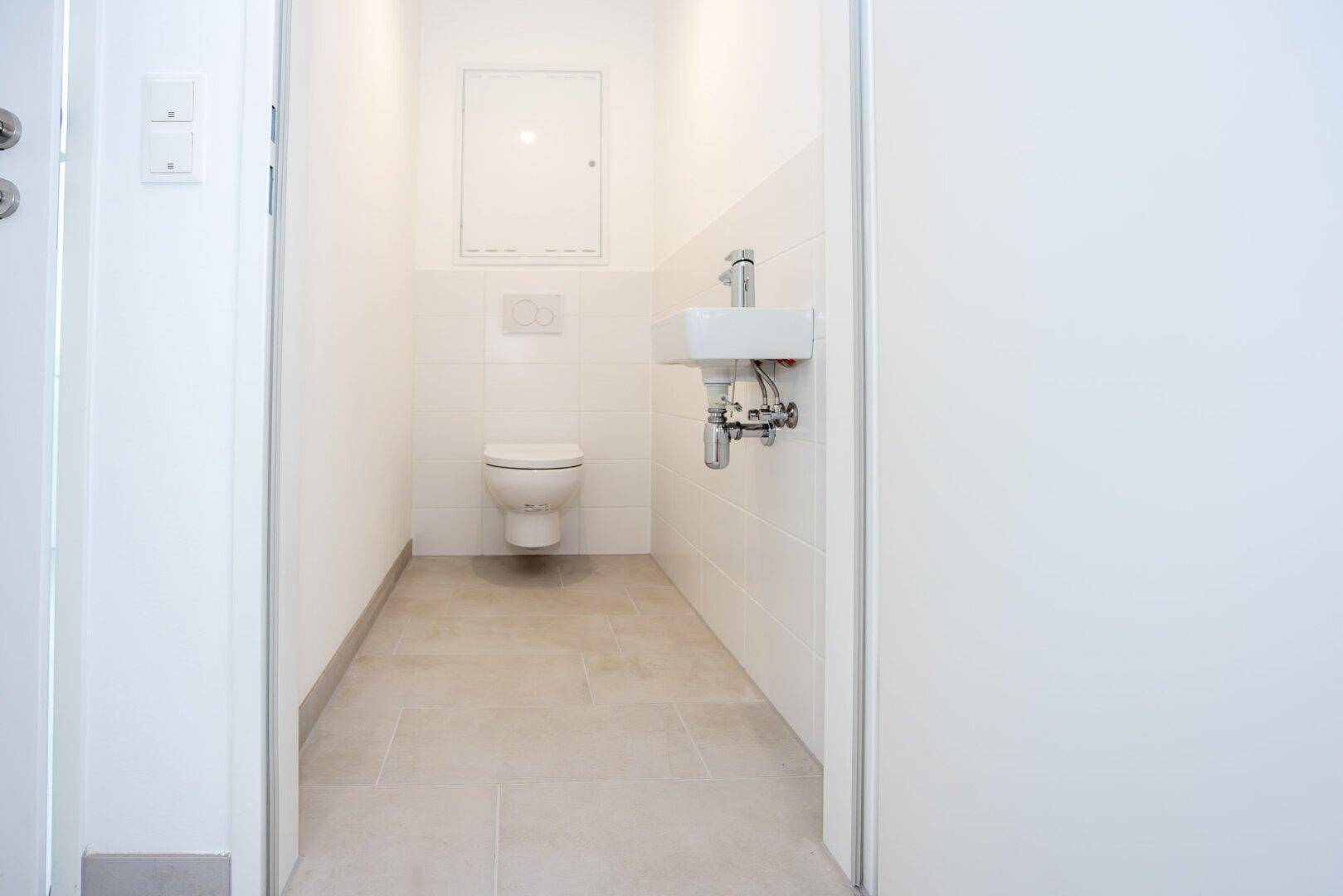 Separates Gäste-WC mit wandhängender Toilette und kleinem Waschbecken, in hellen Fliesen gehalten.